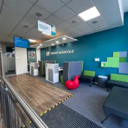 Wnętrze oddziału banku Crédit Agricole z nowoczesną aranżacją: turkusowa ściana, szare i zielone meble, czerwony fotel w kształcie konika i drewniana podłoga z czarno-żółtymi pasami.