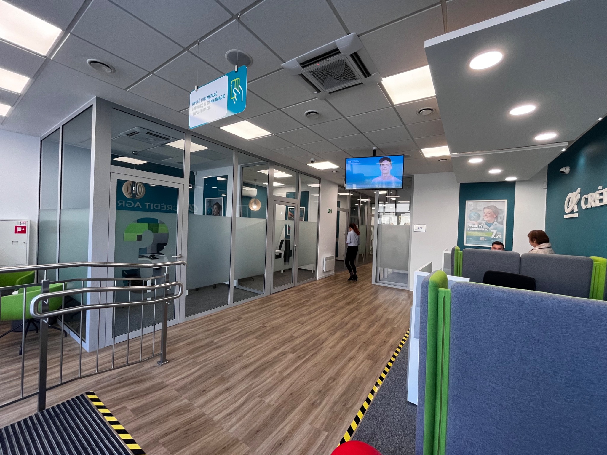 Wnętrze placówki bankowej Credit Agricole z podziałem na strefy obsługi klienta, przeszklone boksy, drewnopodobna podłoga, oświetlenie sufitowe i logo banku na ścianie.