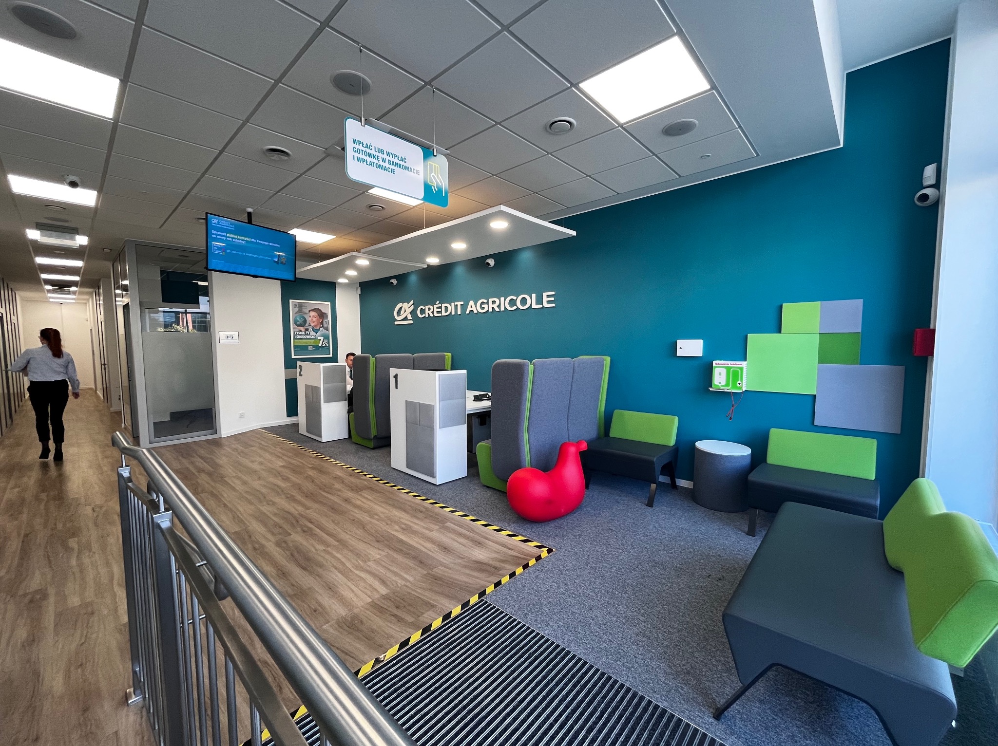 Wnętrze oddziału banku Crédit Agricole z nowoczesną aranżacją: turkusowa ściana, szare i zielone meble, czerwony fotel w kształcie konika i drewniana podłoga z czarno-żółtymi pasami.