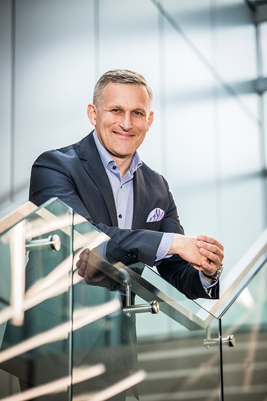 Portret biznesowy uśmiechniętego mężczyzny w garniturze opierającego się o szklaną balustradę schodów w nowoczesnym biurowcu, z chusteczką w kieszonce i zegarkiem na ręku.