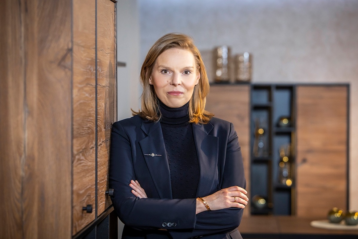 Portret kobiety w granatowym garniturze i czarnym golfie, stojącej na tle drewnianej zabudowy wnętrza z dekoracyjnymi elementami.