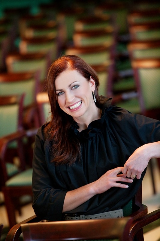 Uśmiechnięta kobieta z rudymi włosami w czarnej bluzce siedzi na krześle na tle rzędów krzeseł.