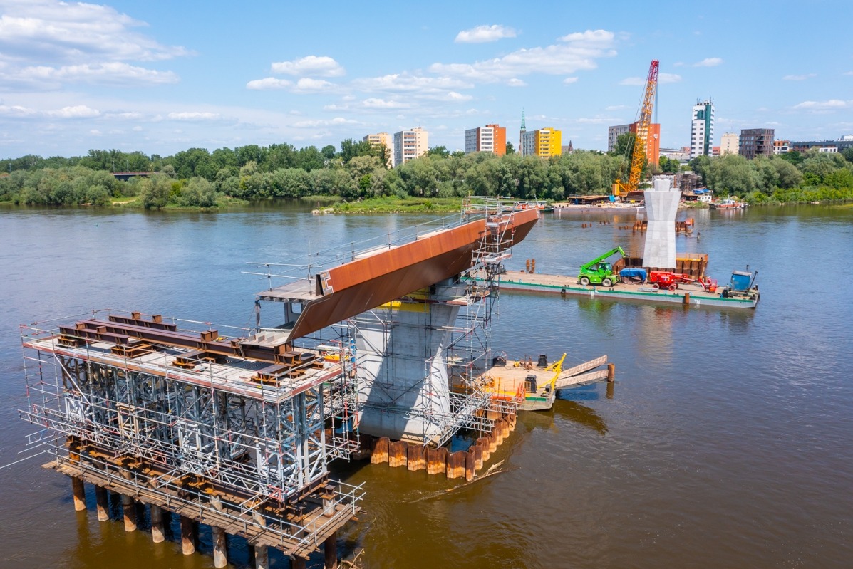 Budowa mostu na rzece, widoczne fragmenty konstrukcji, rusztowania, barki z dźwigami i sprzętem budowlanym, w tle zabudowa miejska.