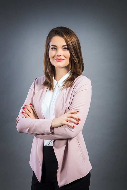 Profesjonalny portret kobiety w różowym żakiecie i białej koszuli, z założonymi rękami na tle szarego studia. Uśmiech, zadbane włosy i czerwone paznokcie.