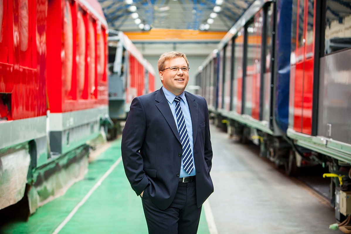 Mężczyzna w garniturze, z rękami w kieszeniach, stoi w hali z wagonami kolejowymi. Uśmiechnięty, patrzy w górę. Ujęcie z dołu, symetryczny kadr.