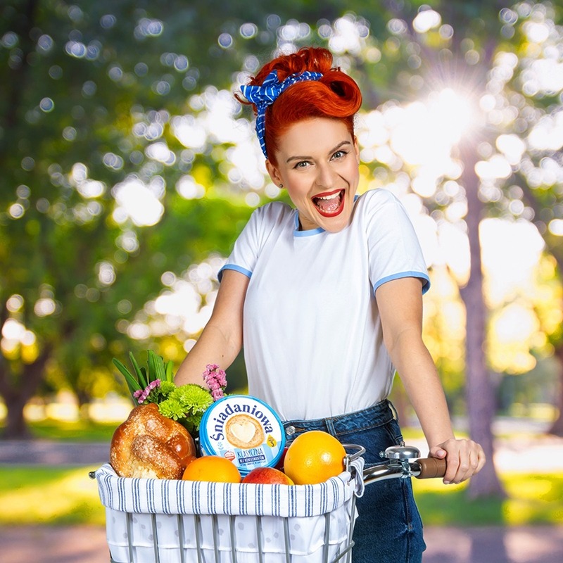 Uśmiechnięta kobieta z czerwonymi włosami na rowerze, w koszyku jedzenie i ser 'Śniadaniowa', w tle zieleń parku i słońce.