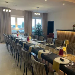 Elegancko nakryty stół na przyjęcie świąteczne z ciemnym obrusem, złotym bieżnikiem i dekoracjami, w tle choinka i bar z podświetleniem.