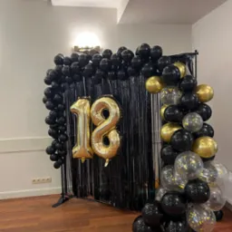 Dekoracja na osiemnaste urodziny: złote balony w kształcie cyfr '18' na tle czarnej, błyszczącej kurtyny, otoczone girlandą z czarnych, złotych i przezroczystych balonów z konfetti.