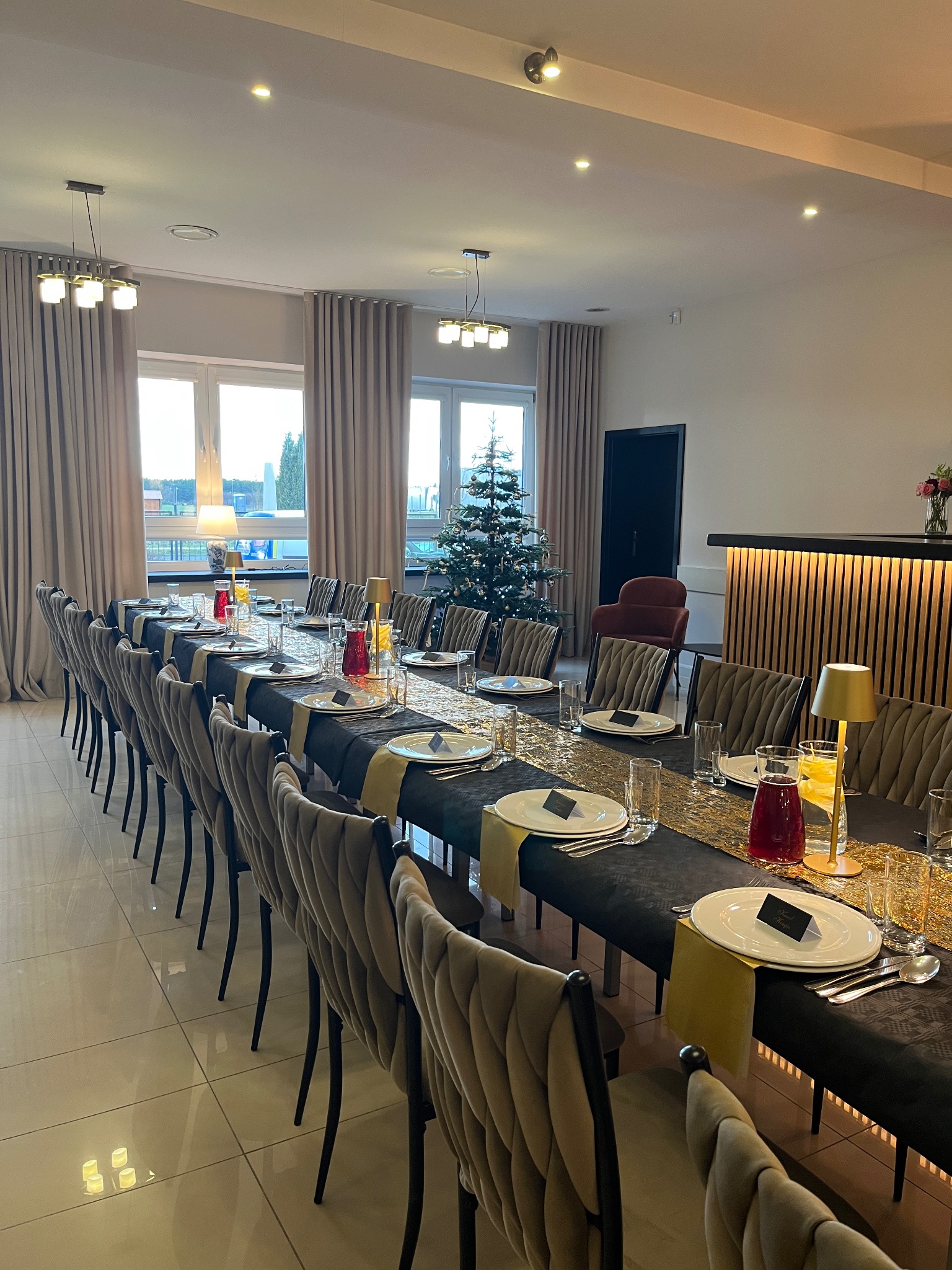 Elegancko nakryty stół na przyjęcie świąteczne z ciemnym obrusem, złotym bieżnikiem i dekoracjami, w tle choinka i bar z podświetleniem.