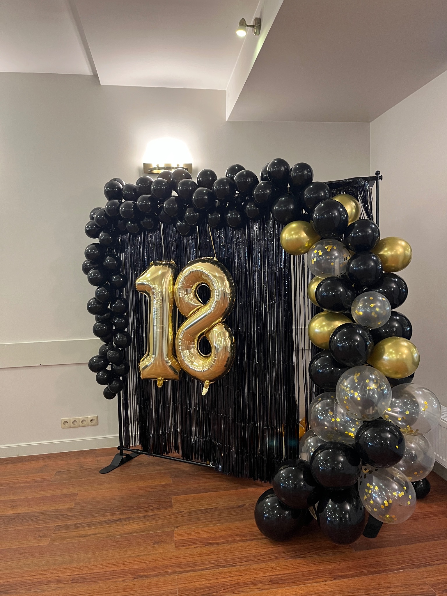 Dekoracja na osiemnaste urodziny: złote balony w kształcie cyfr '18' na tle czarnej, błyszczącej kurtyny, otoczone girlandą z czarnych, złotych i przezroczystych balonów z konfetti.