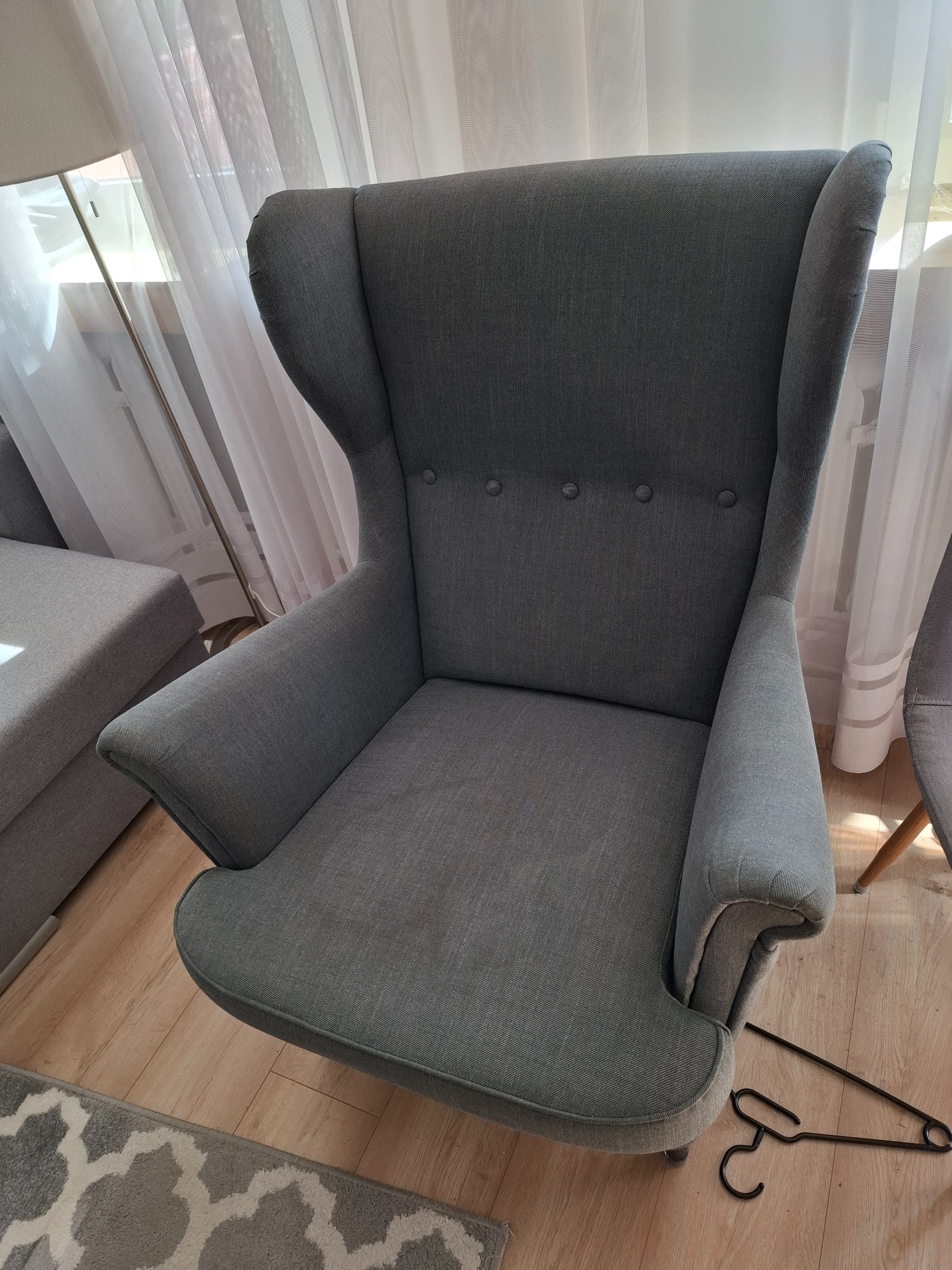 Szary fotel uszak na tle okna z firanką. Na podłodze leży metalowy wieszak. Widoczny fragment sofy i dywanu w odcieniach szarości. Delikatne światło dzienne.