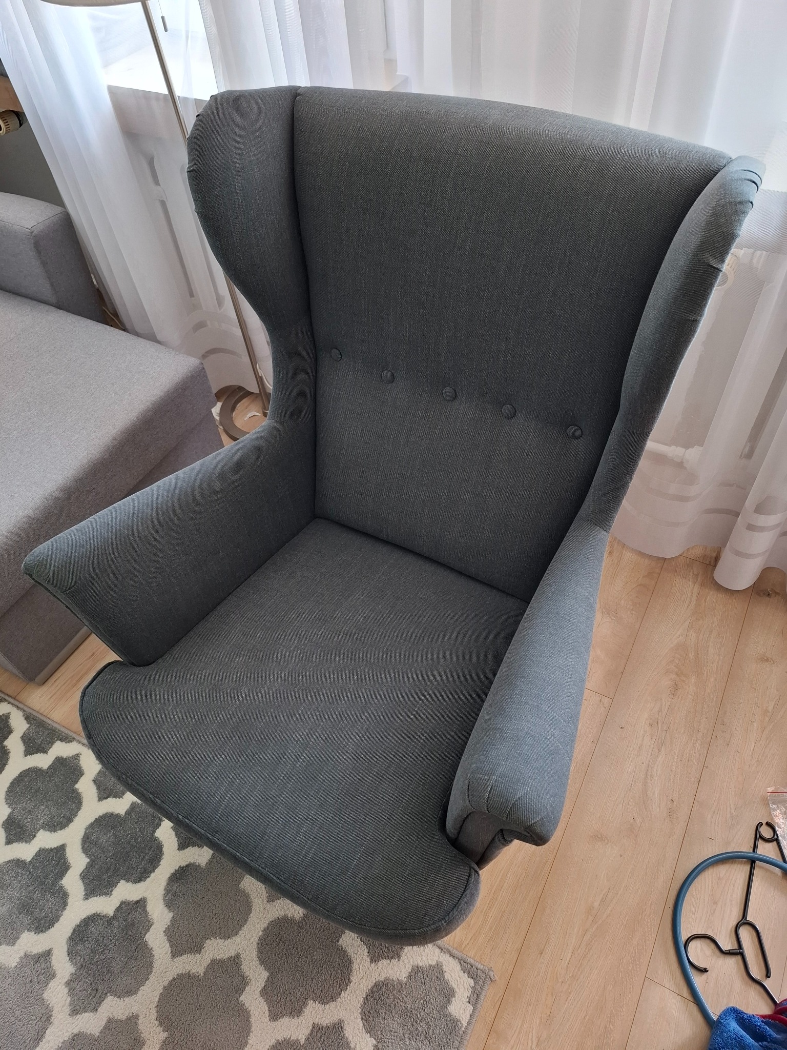 Szary fotel uszak z tapicerowanym siedziskiem na tle jasnych zasłon i wzorzystego dywanu. Widok z góry, podkreślający kształt i fakturę tkaniny.