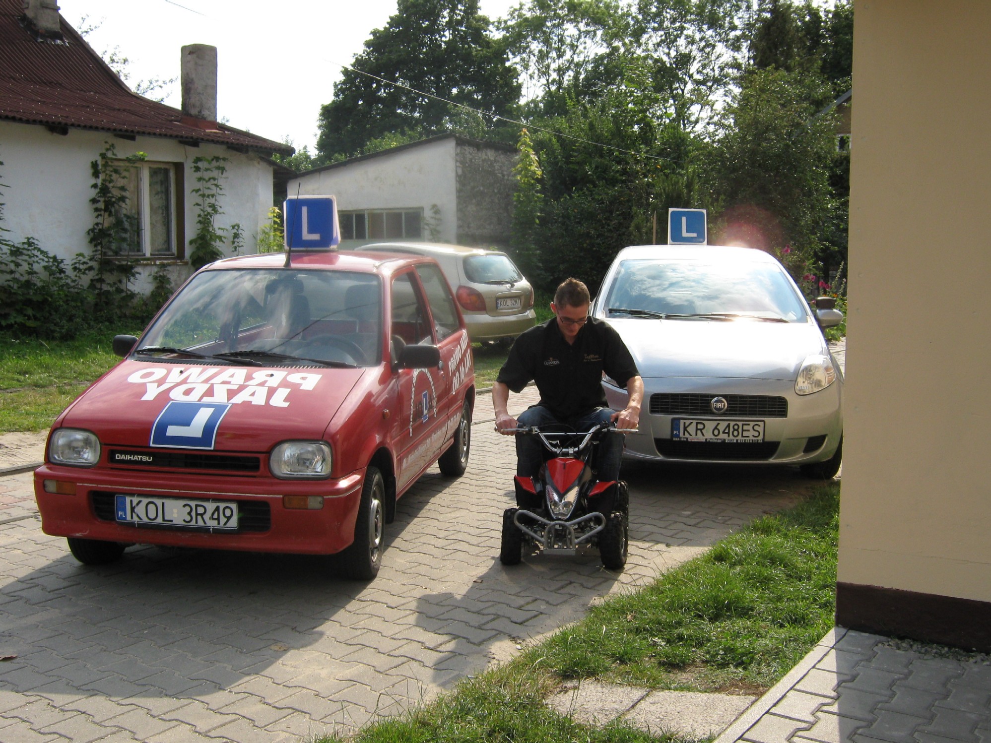 Mężczyzna na quadzie pomiędzy dwoma samochodami z literą 'L' na dachu, symbolizującymi naukę jazdy.