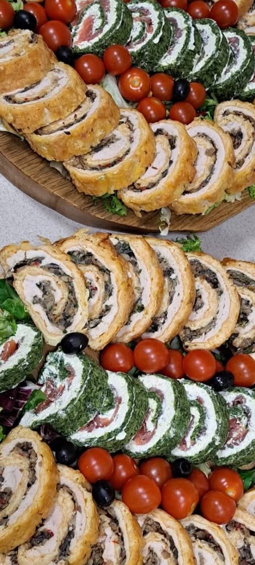 Dekoracyjna taca z roladkami mięsnymi i warzywnymi, udekorowana pomidorkami koktajlowymi i oliwkami, prezentująca różnorodność smaków i estetyczne podanie.