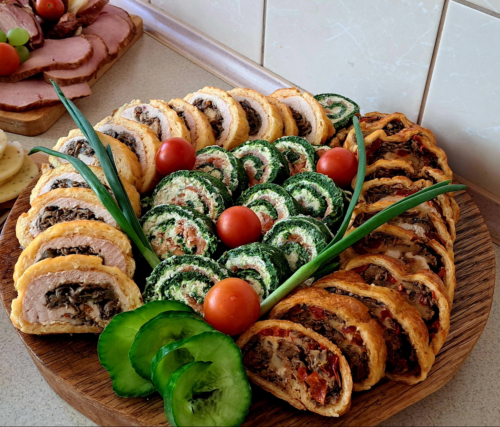 Elegancko ułożona na drewnianej desce kompozycja z rolad mięsnych, szpinakowych i z ciasta francuskiego, udekorowana pomidorkami i ogórkiem, gotowa do serwowania.