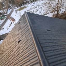 BUDRAF - Nowy, ciemny dach z dachówki ceramicznej, widok z góry na fragment połaci dachowej z zamontowanym gąsiorem, w tle ośnieżony teren i drzewa.
