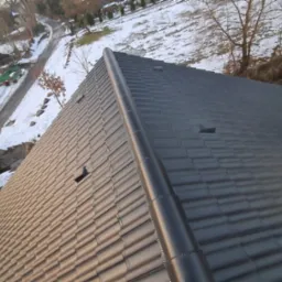 Nowy, ciemny dach z dachówki ceramicznej, widok z góry na fragment połaci dachowej z zamontowanym gąsiorem, w tle ośnieżony teren i drzewa.