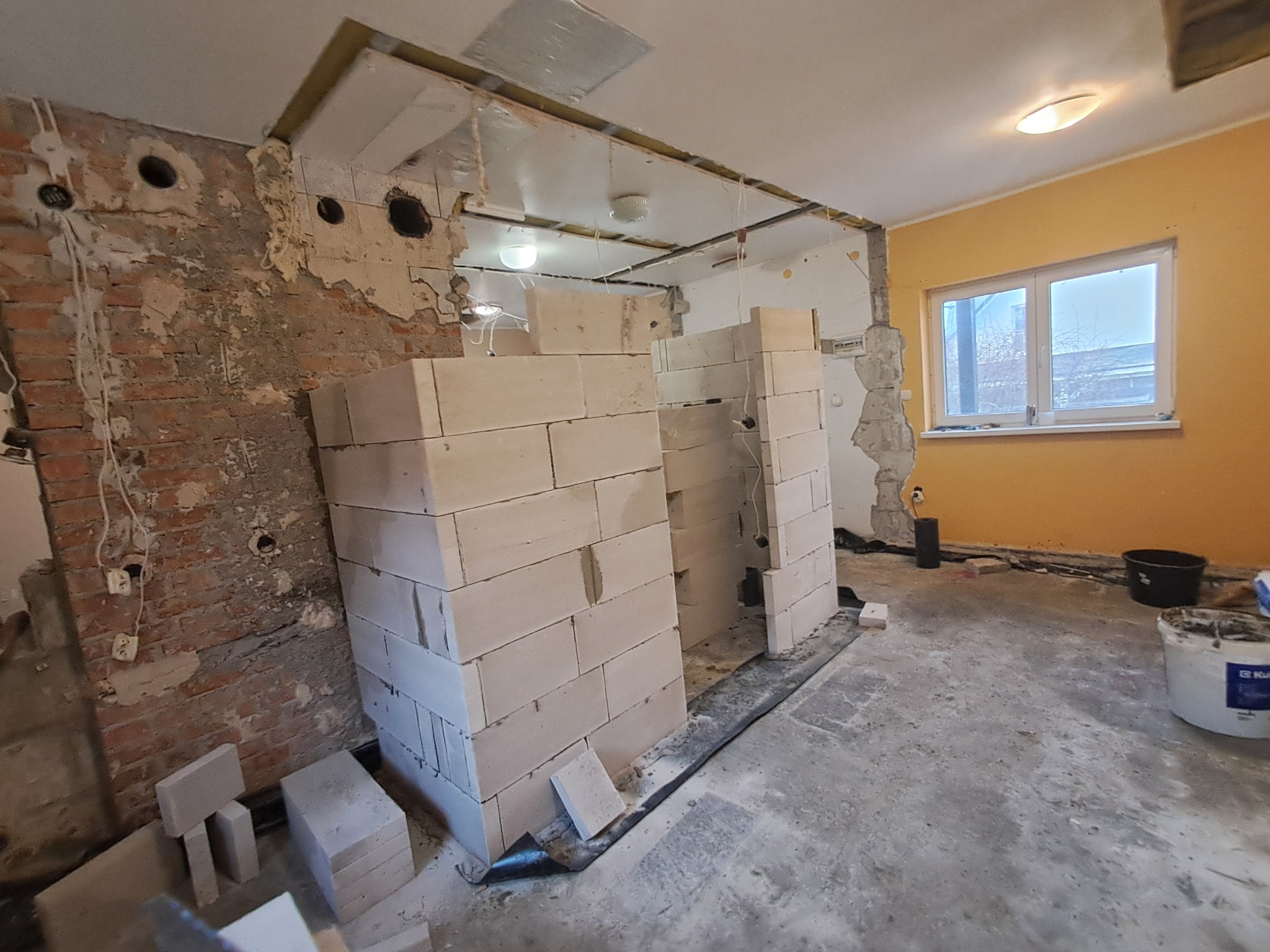 Wnętrze w trakcie remontu: surowa cegła, częściowo otynkowane ściany, murowane ścianki działowe z bloczków, widoczne instalacje elektryczne, sufit z odsłoniętą konstrukcją, okno w żółtej ścianie.