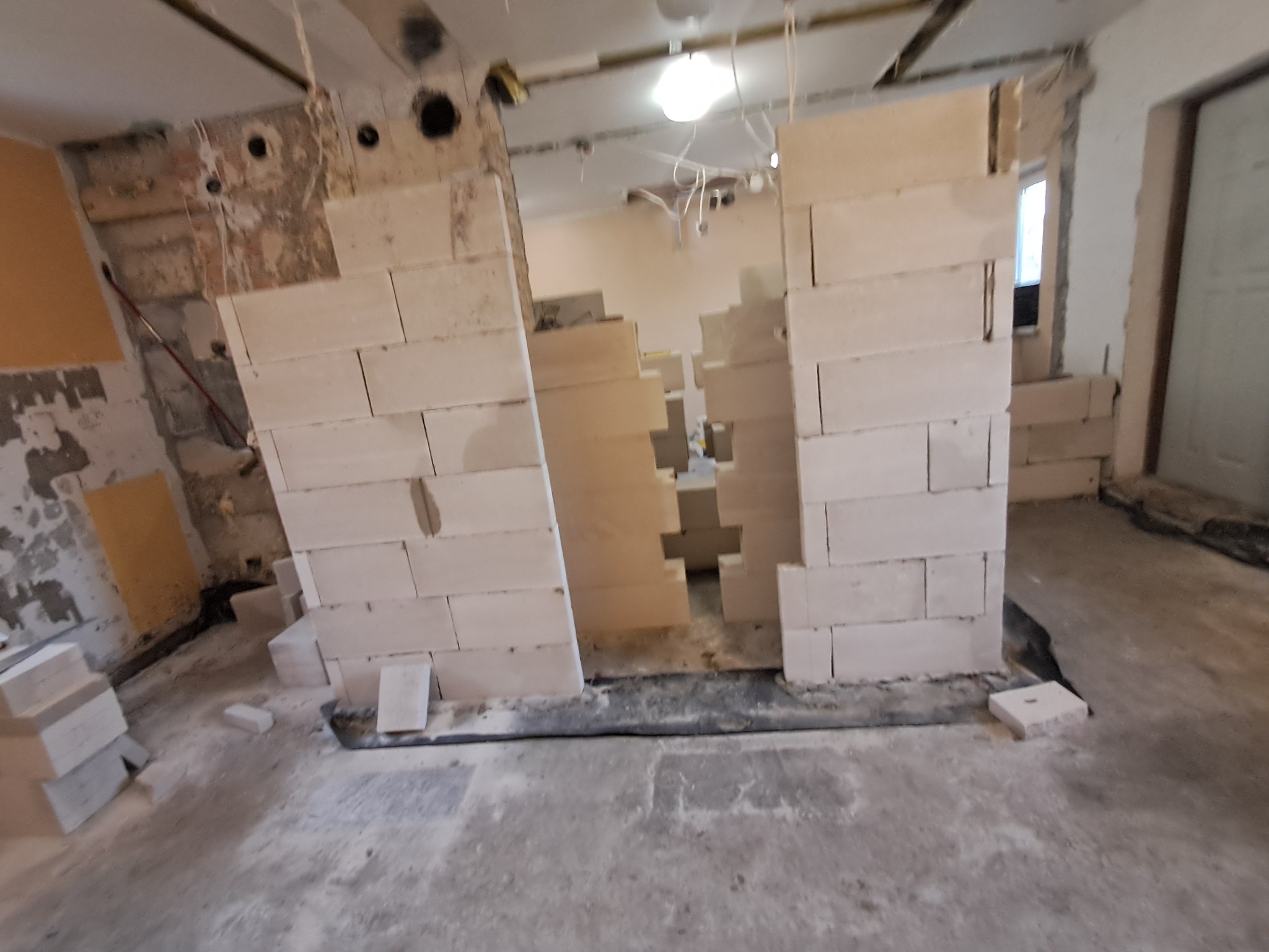 Wnętrze w trakcie budowy z fragmentami ścian z białych bloczków, widoczne instalacje pod sufitem i surowe wykończenie ścian.