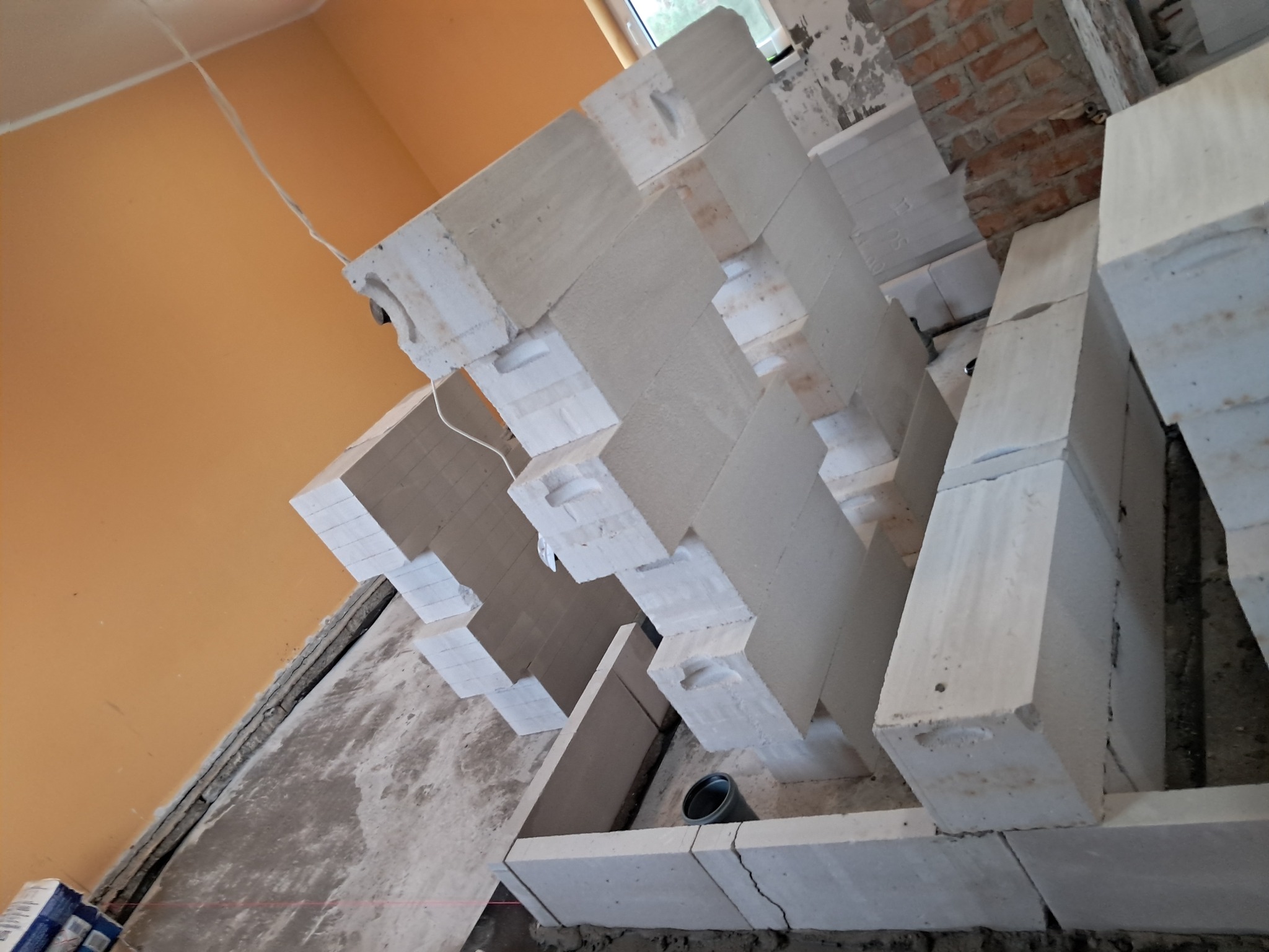 Wznoszenie komina z białych bloczków ceramicznych, widoczne elementy konstrukcyjne, ślady zaprawy i wystające przewody elektryczne na tle surowej ściany i podłogi.