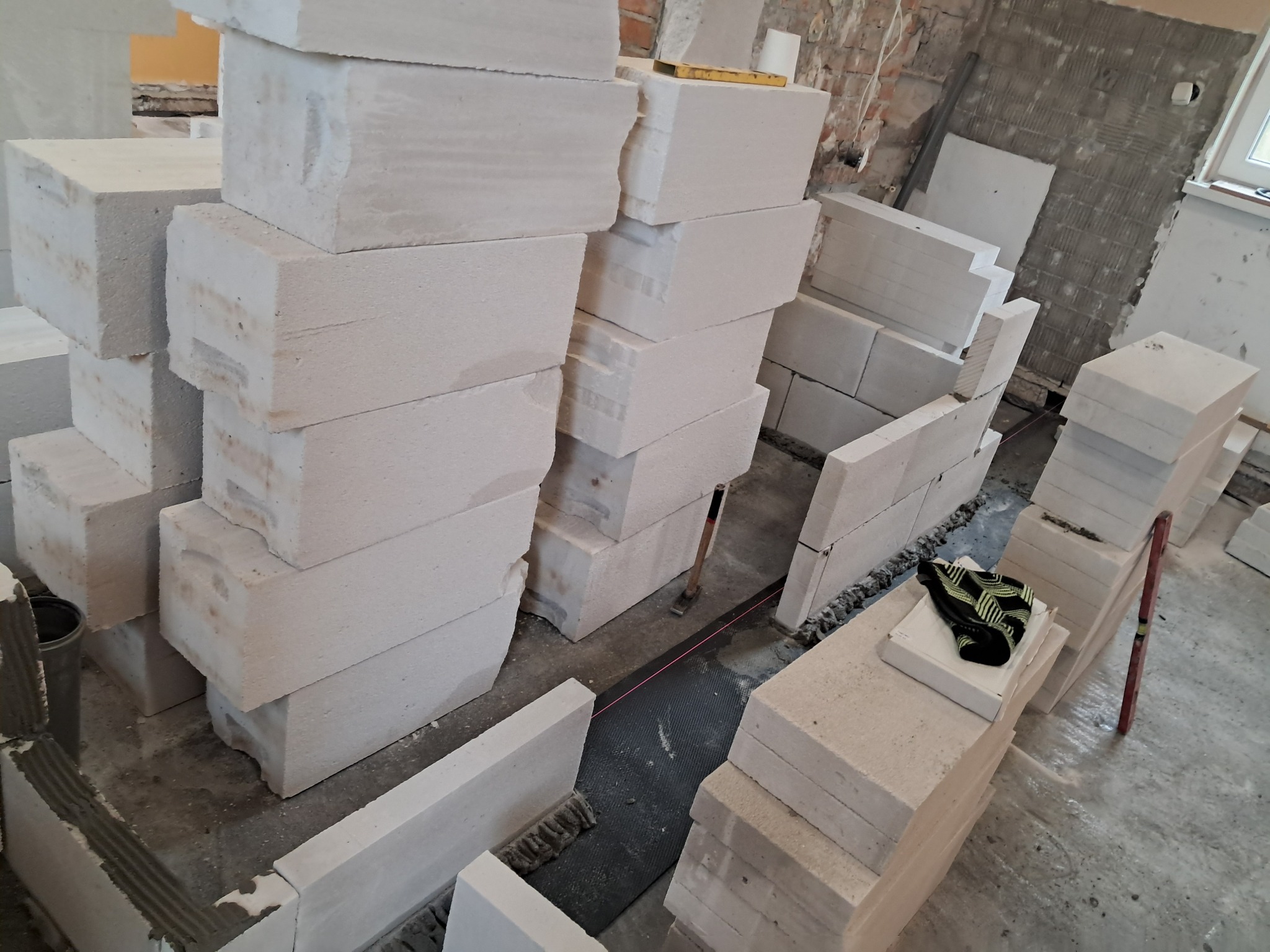 Budowa ściany z białych bloczków betonu komórkowego, widoczne narzędzia murarskie, poziomica i sznurek traserski, surowe ściany w tle.