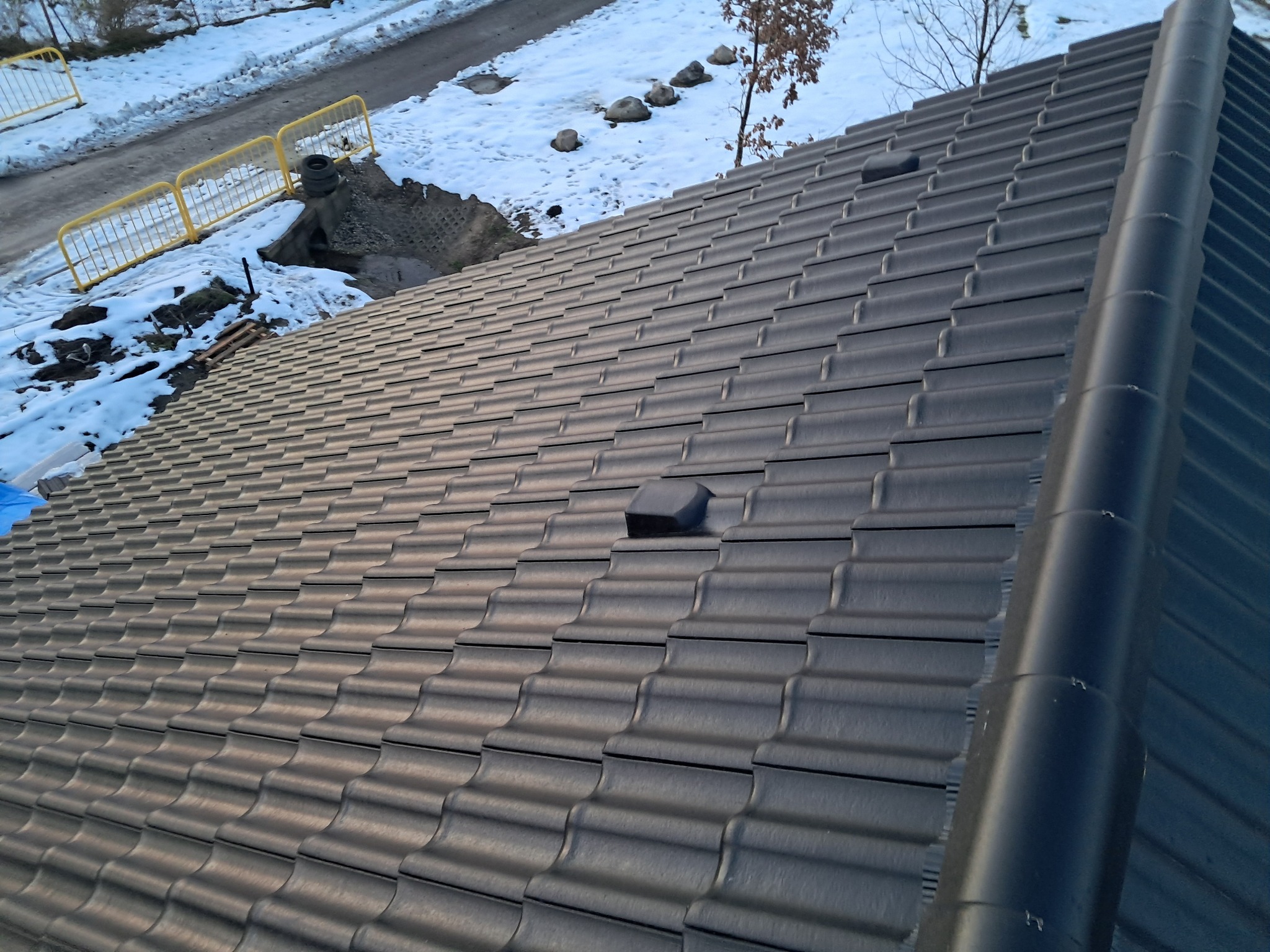 Ukośny widok na nowy, grafitowy dach z ceramicznej dachówki falistej, z widocznym kominkiem wentylacyjnym i fragmentem rynny, na tle ośnieżonego otoczenia.