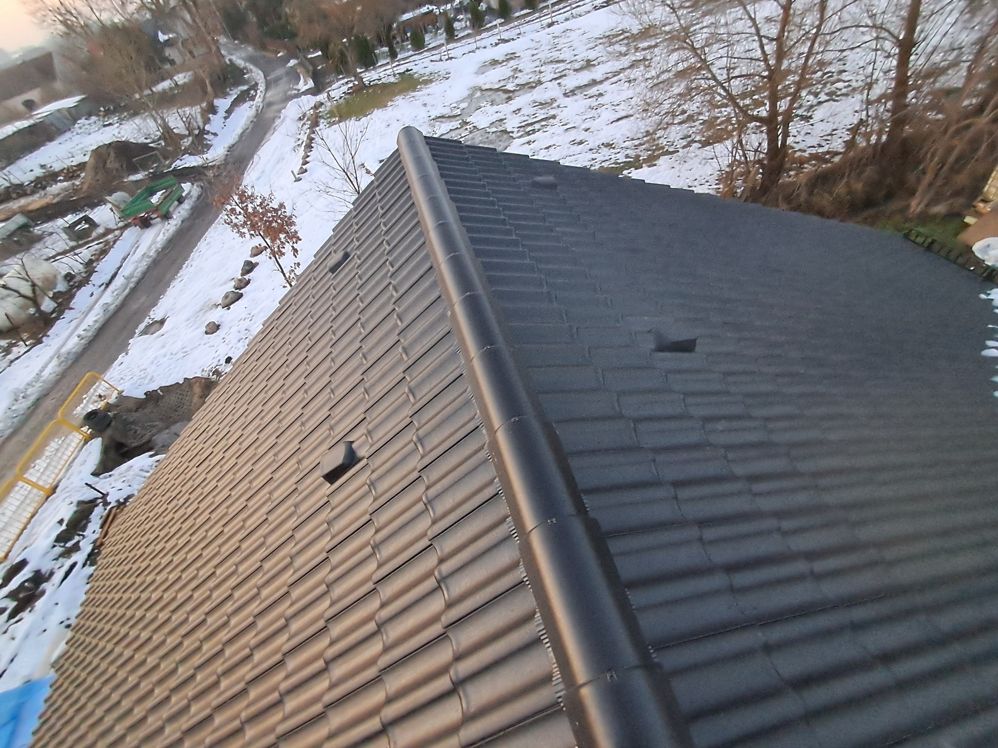 Nowy, ciemny dach z dachówki ceramicznej, widok z góry na fragment połaci dachowej z zamontowanym gąsiorem, w tle ośnieżony teren i drzewa.