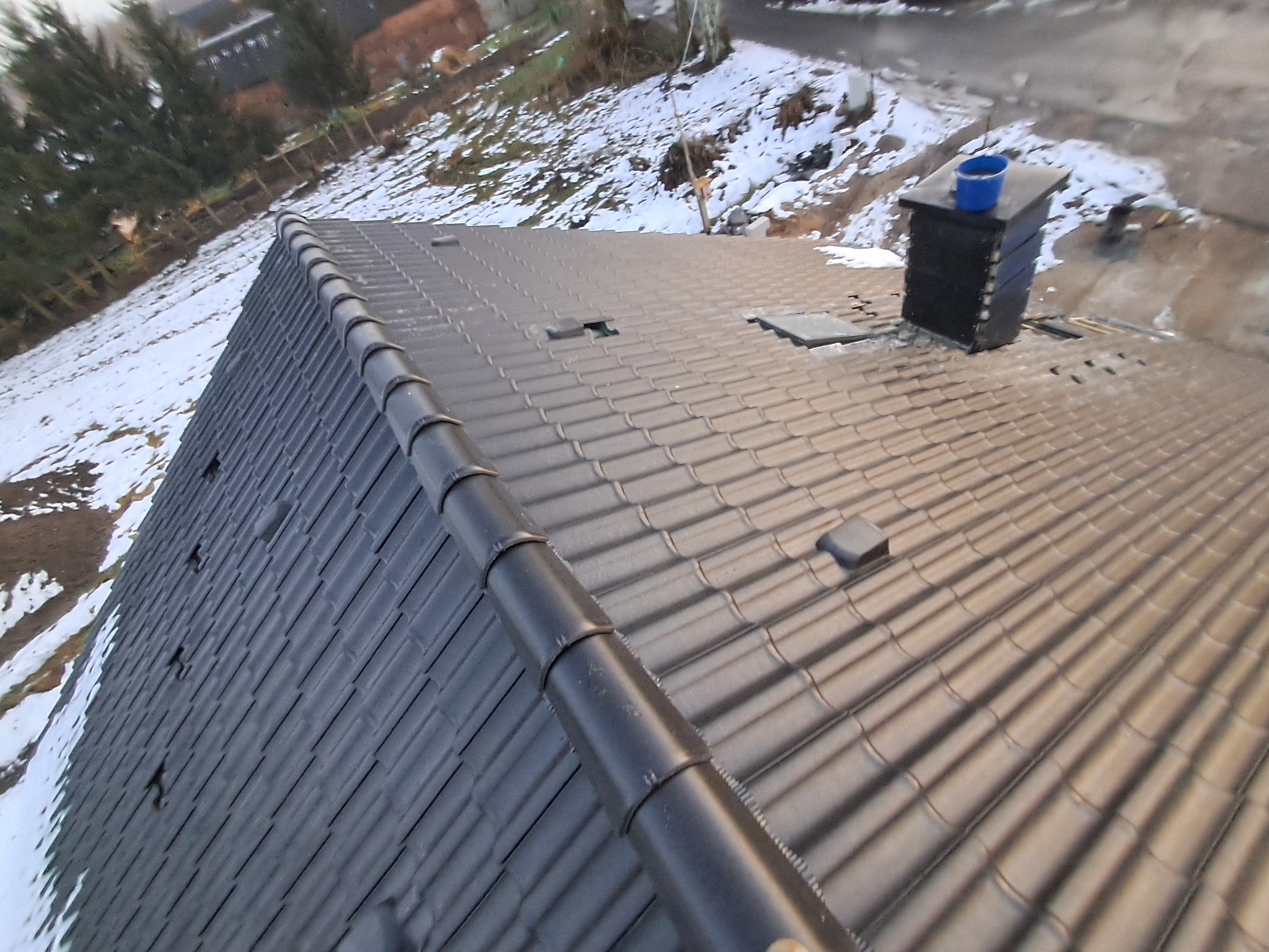 Szary dach z dachówki ceramicznej widziany z góry, z fragmentami śniegu na dachu i w tle, oraz niebieskie wiadro na kominie.