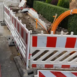 Oznakowany wykop wzdłuż chodnika z widocznymi pomarańczowymi kablami i taczką w tle.