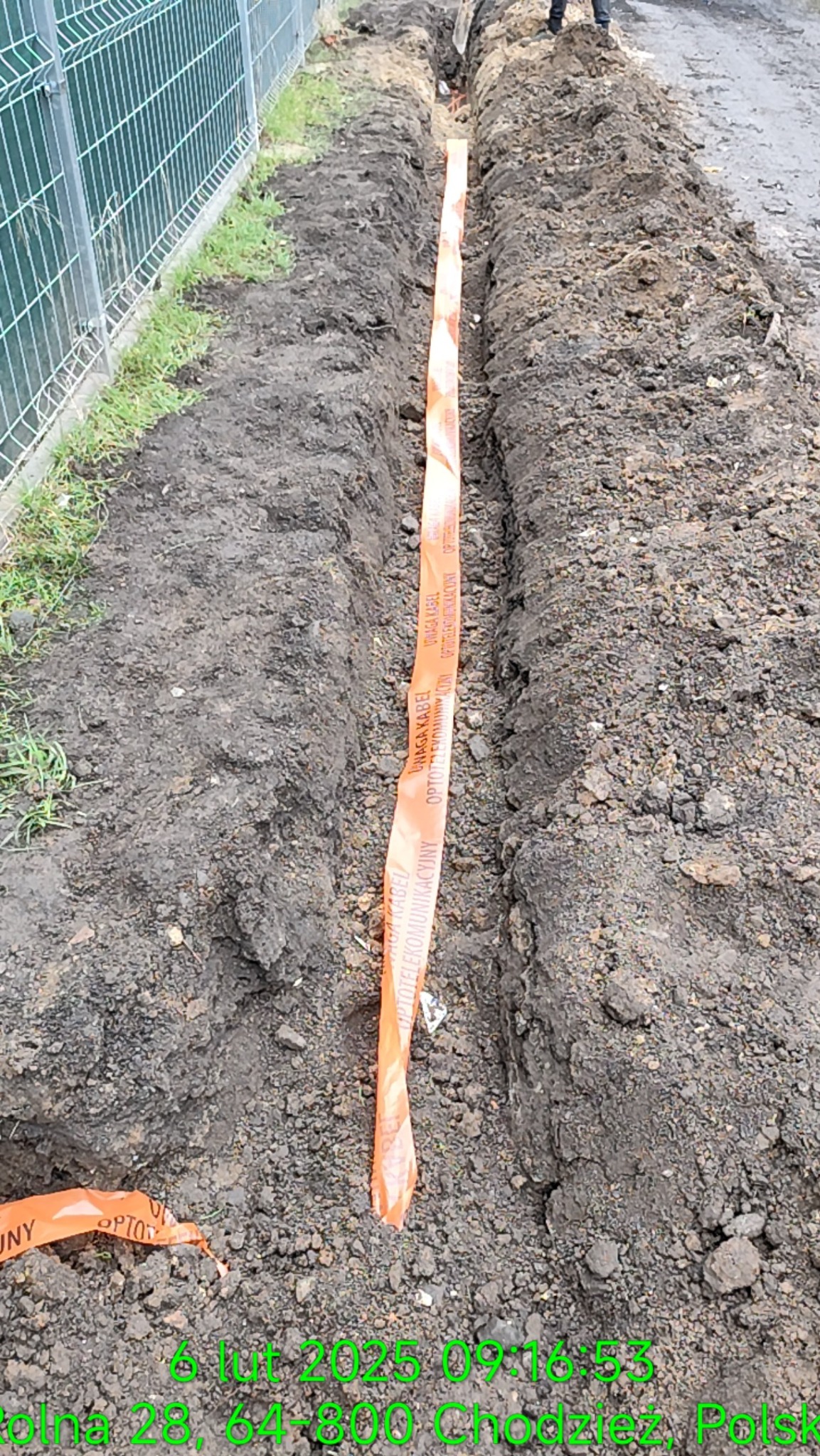 Wąski wykop w ziemi, zabezpieczony pomarańczową taśmą ostrzegawczą z napisem 'Uwaga kabel optotelekomunikacyjny', widoczny fragment ogrodzenia z siatki.