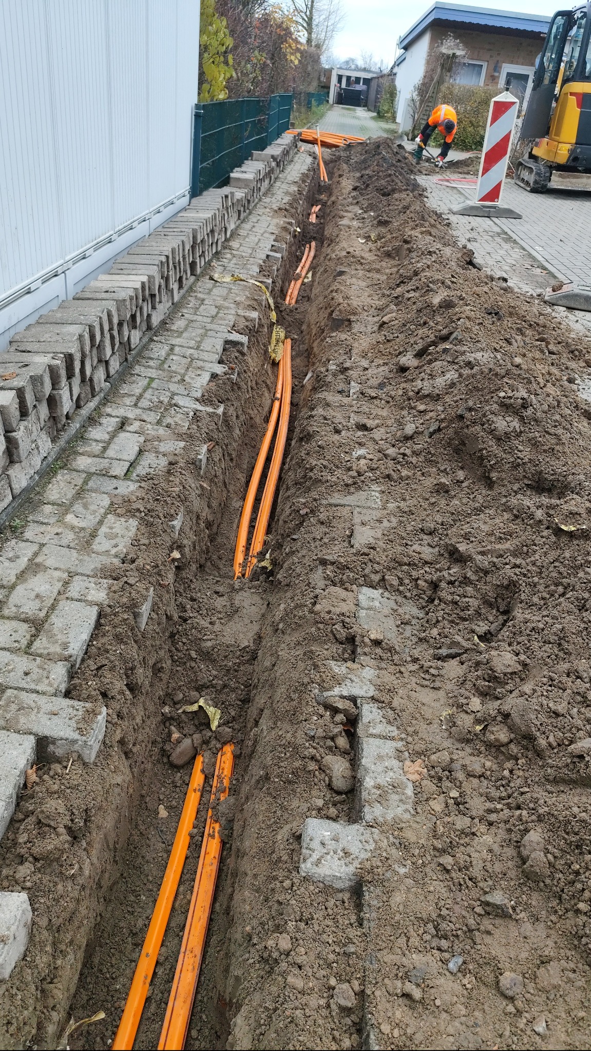 Wykopany rów w ziemi z ułożonymi pomarańczowymi rurami instalacyjnymi, widoczny pracownik w pomarańczowym ubraniu oraz minikoparka w tle.
