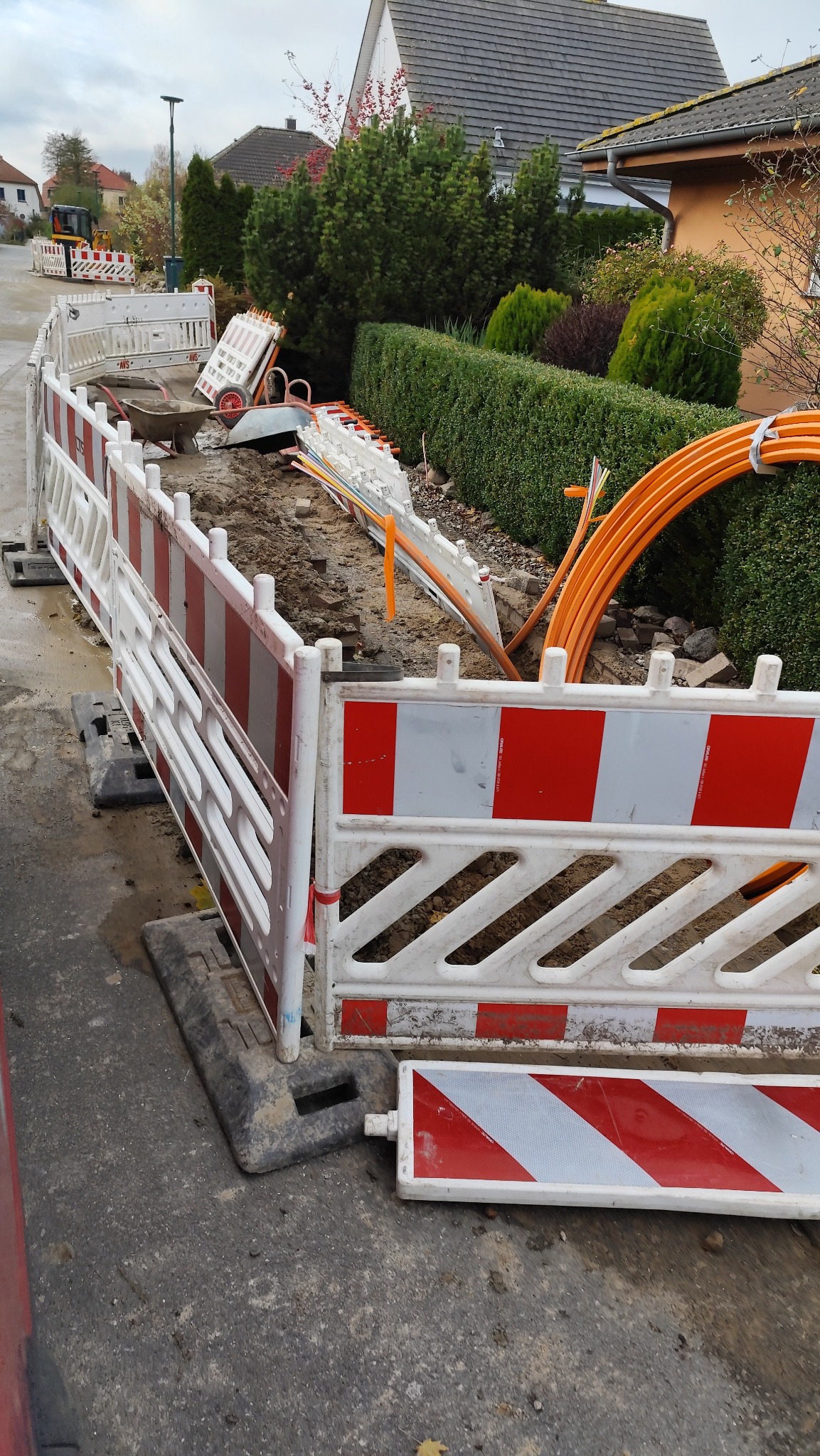 Oznakowany wykop wzdłuż chodnika z widocznymi pomarańczowymi kablami i taczką w tle.