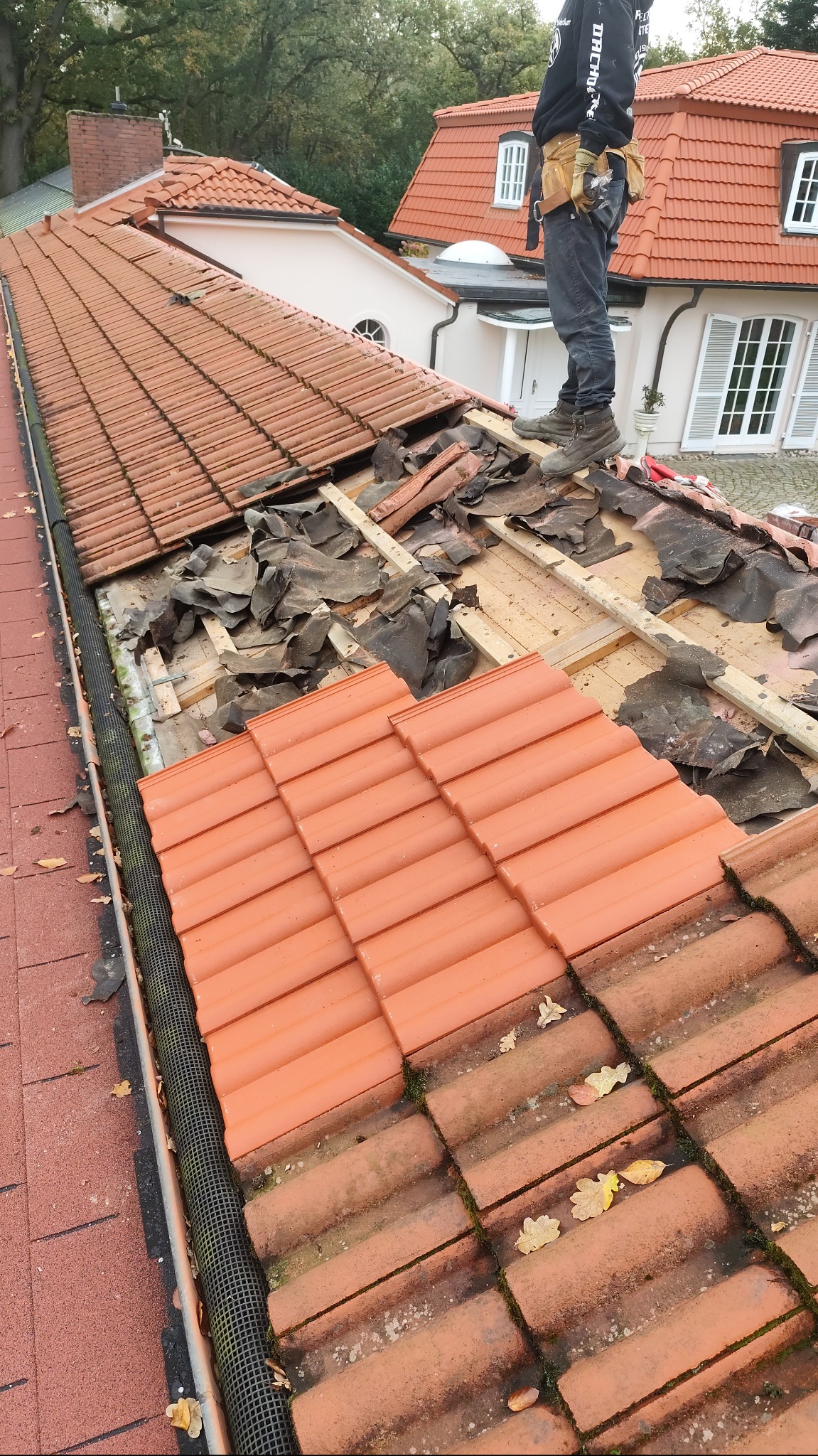 Prace dekarskie na dachu domu z czerwoną dachówką, widoczne uszkodzone fragmenty pokrycia, dekarz w ubraniu roboczym i pasem z narzędziami stoi na dachu.