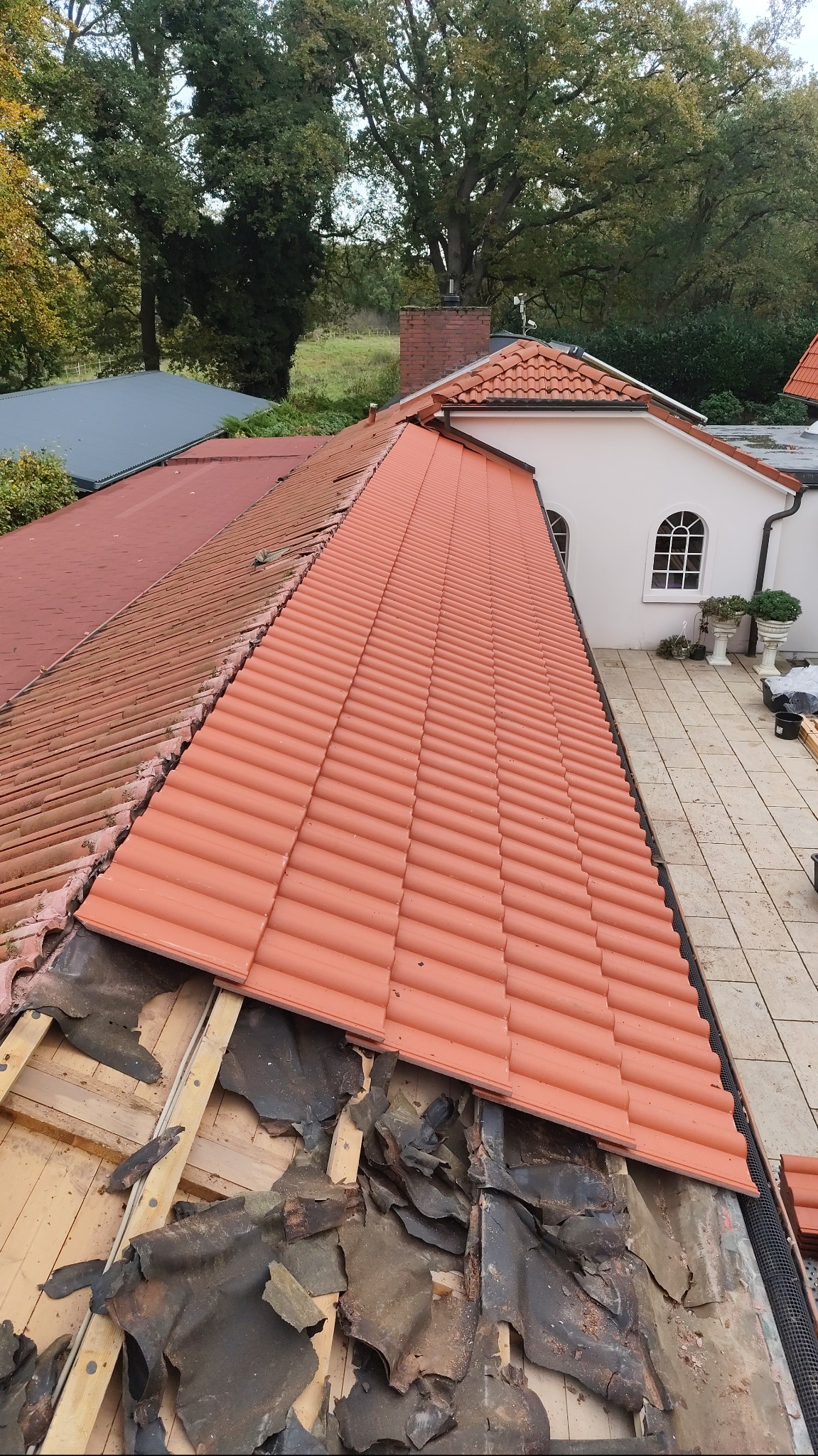 Częściowo zdjęte stare dachówki i nowa, pomarańczowa dachówka ceramiczna na dachu domu jednorodzinnego z widoczną konstrukcją drewnianą i resztkami starej izolacji.