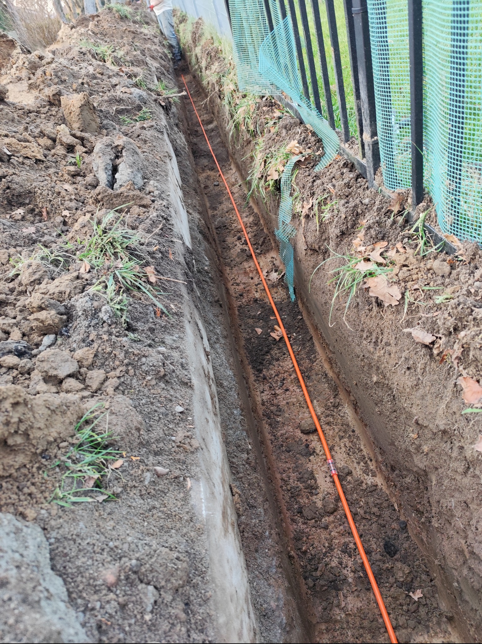 Wąski wykop w ziemi z pomarańczowym przewodem, widoczna granica działki z czarnym metalowym ogrodzeniem i zieloną siatką.