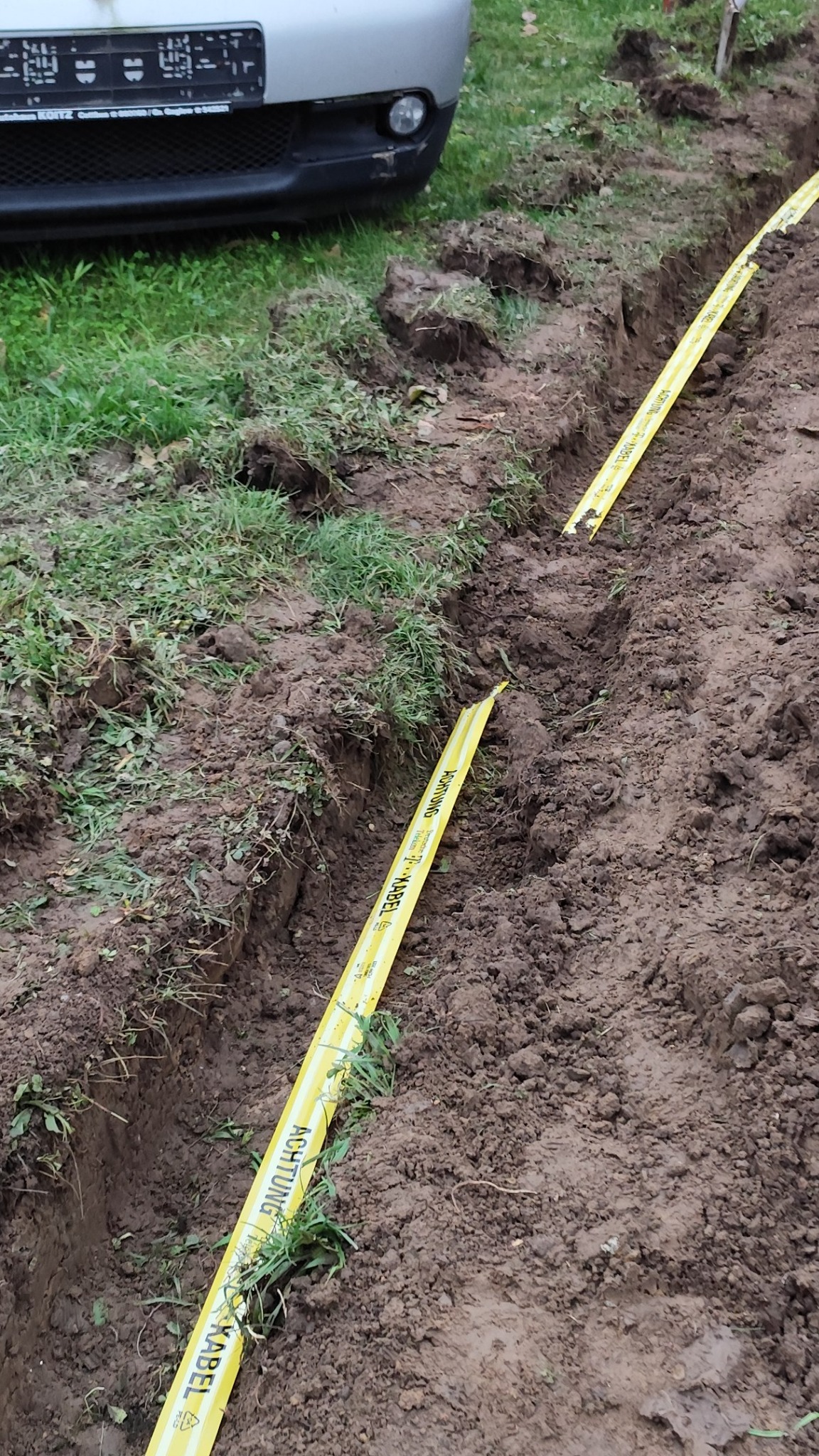 Wąski wykop w ziemi z żółtą taśmą ostrzegawczą 'Achtung Kabel' zabezpieczającą kable, widoczny fragment samochodu w tle.