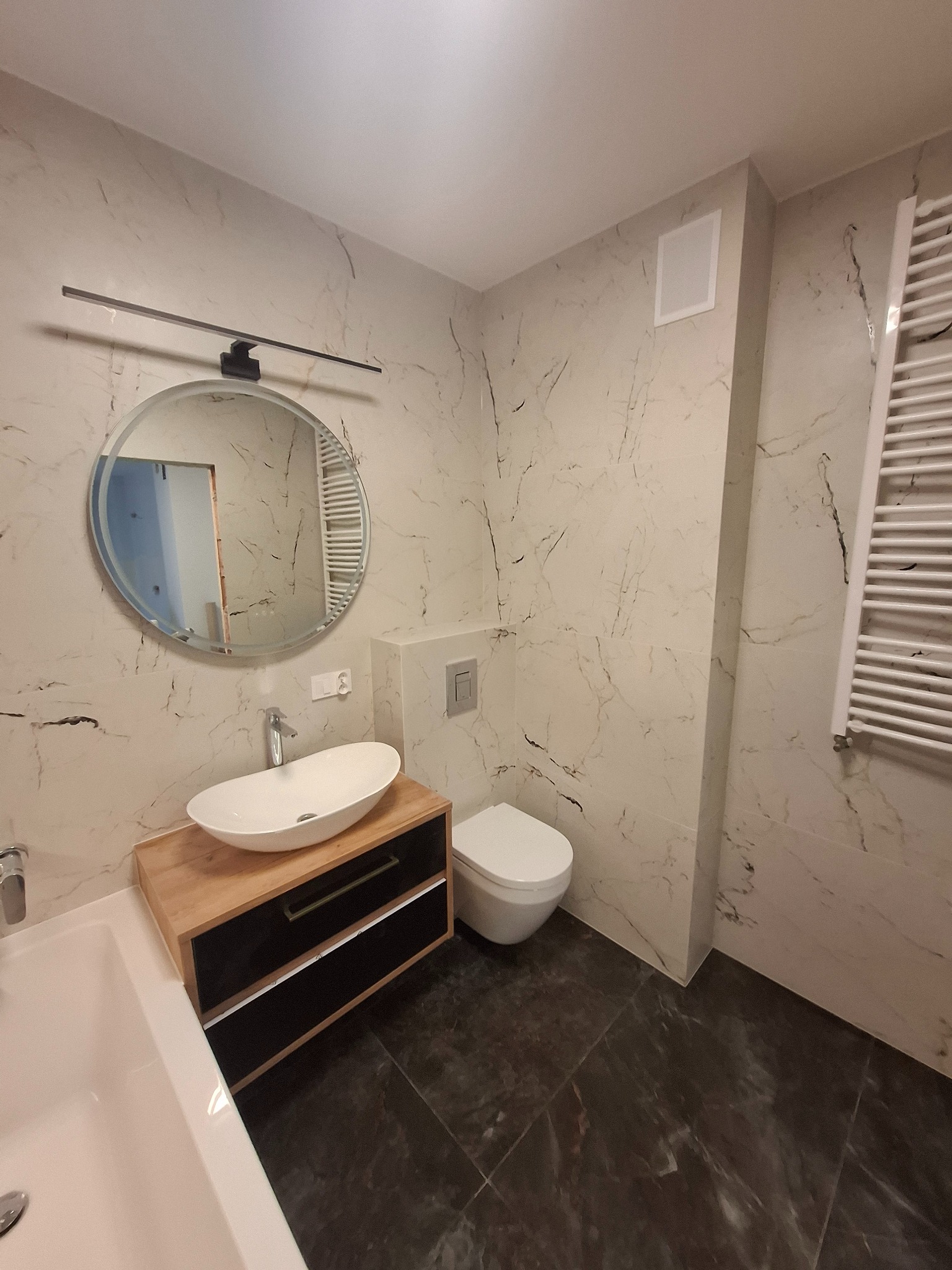 Nowoczesna łazienka z marmurowymi płytkami, okrągłym lustrem z oświetleniem i drewnianą szafką pod umywalką. Grzejnik drabinkowy i toaleta w tle.