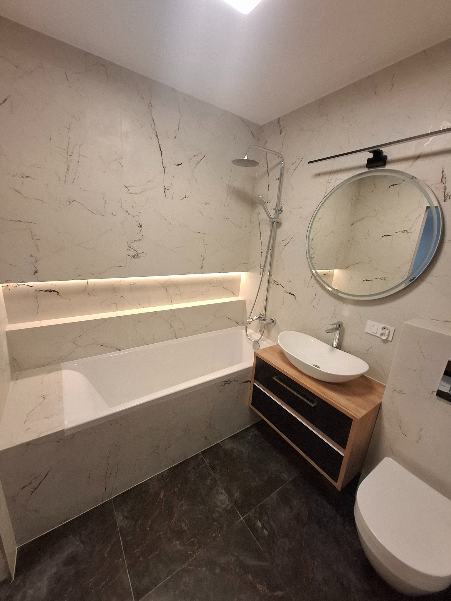 Nowoczesna łazienka z marmurowymi płytkami, podświetlaną wnęką nad wanną, okrągłym lustrem i ciemną podłogą. Umywalka nablatowa na szafce z drewnianym blatem.