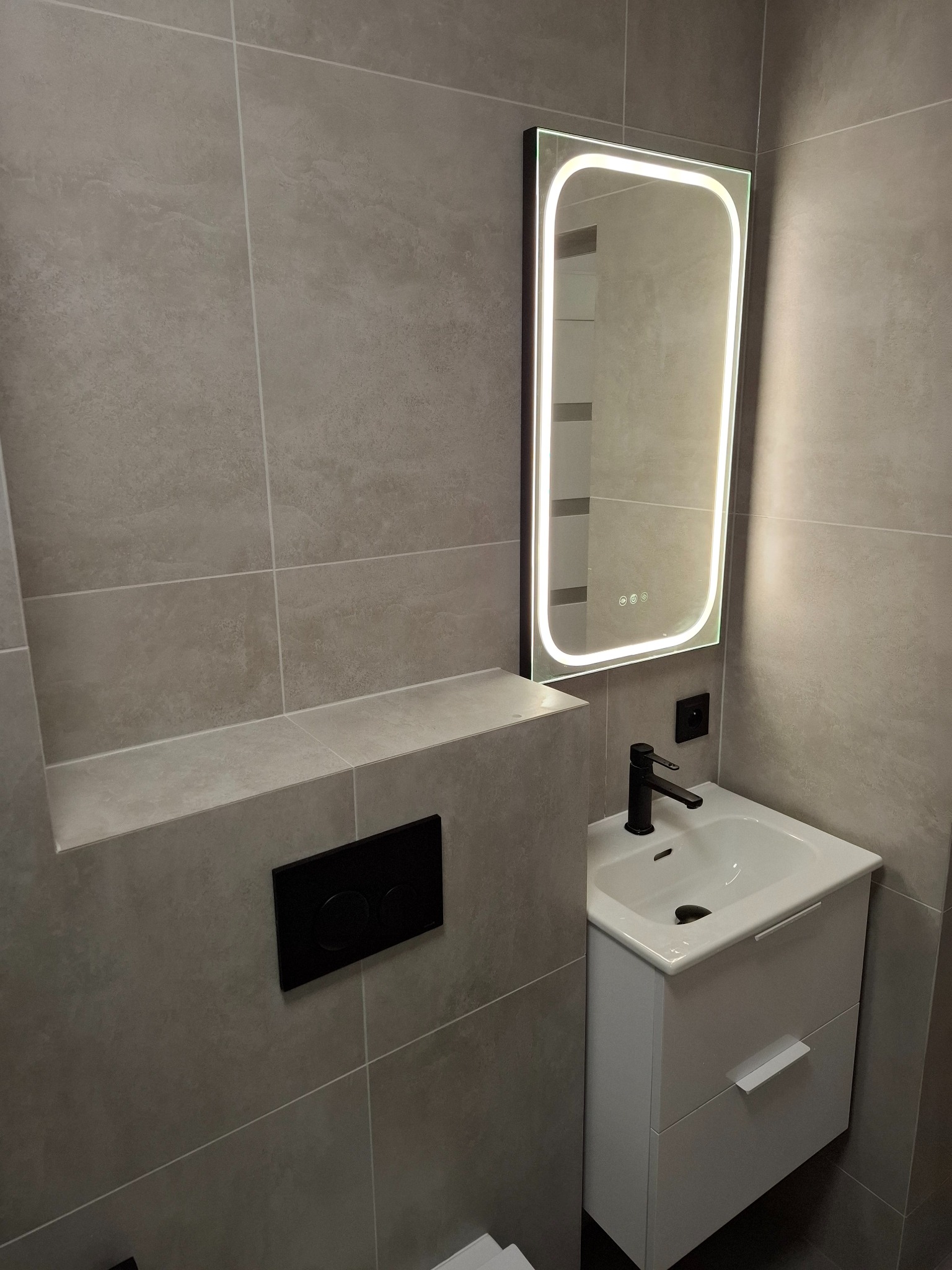 Nowoczesna łazienka w Warszawie: szare płytki, podświetlane lustro, czarna armatura i biała szafka pod umywalką. Minimalistyczny design.