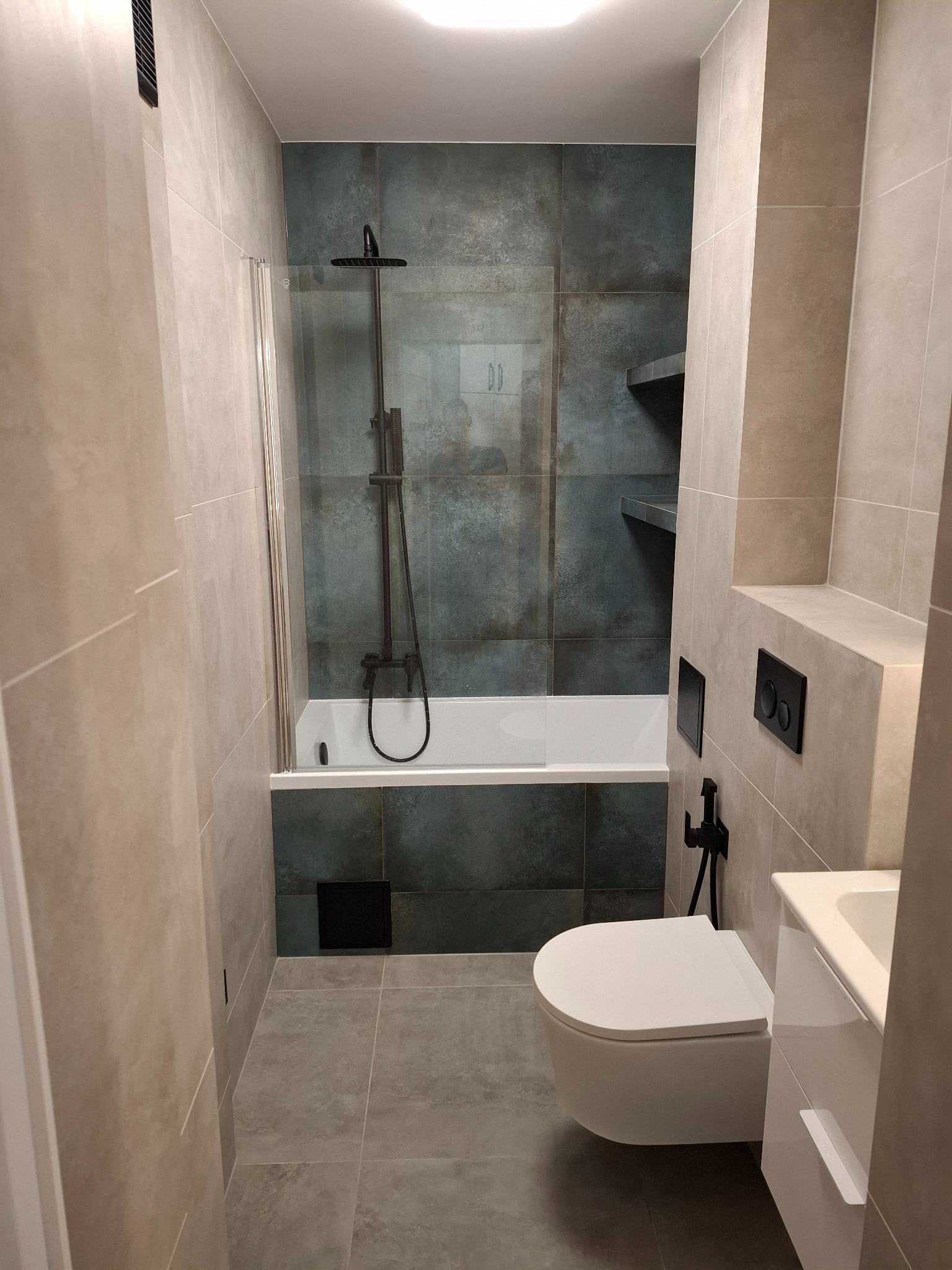 Nowoczesna łazienka z wanną za szklanym parawanem, wiszącą toaletą i umywalką. Ciemne płytki imitujące beton kontrastują z jasnymi płytkami na ścianach.
