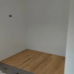 Fragment pokoju w trakcie prac wykończeniowych: częściowo ułożona podłoga z paneli drewnopodobnych, widoczna mata pod panele z nadrukowanym logo producenta, przygotowane arkusze folii ochronnej...