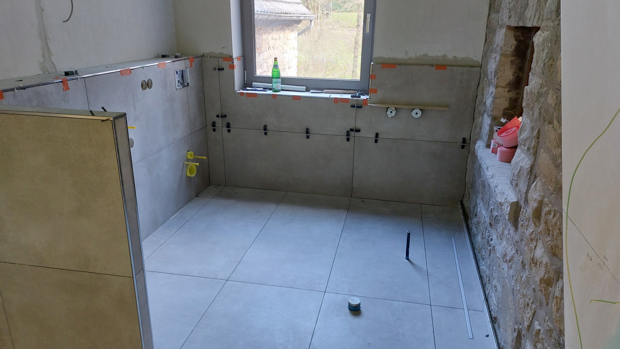 Łazienka w trakcie remontu: szare płytki ułożone na podłodze i ścianach, widoczne krzyżyki dystansowe, butelka wody na parapecie okna, ściana z kamienia z wnęką.