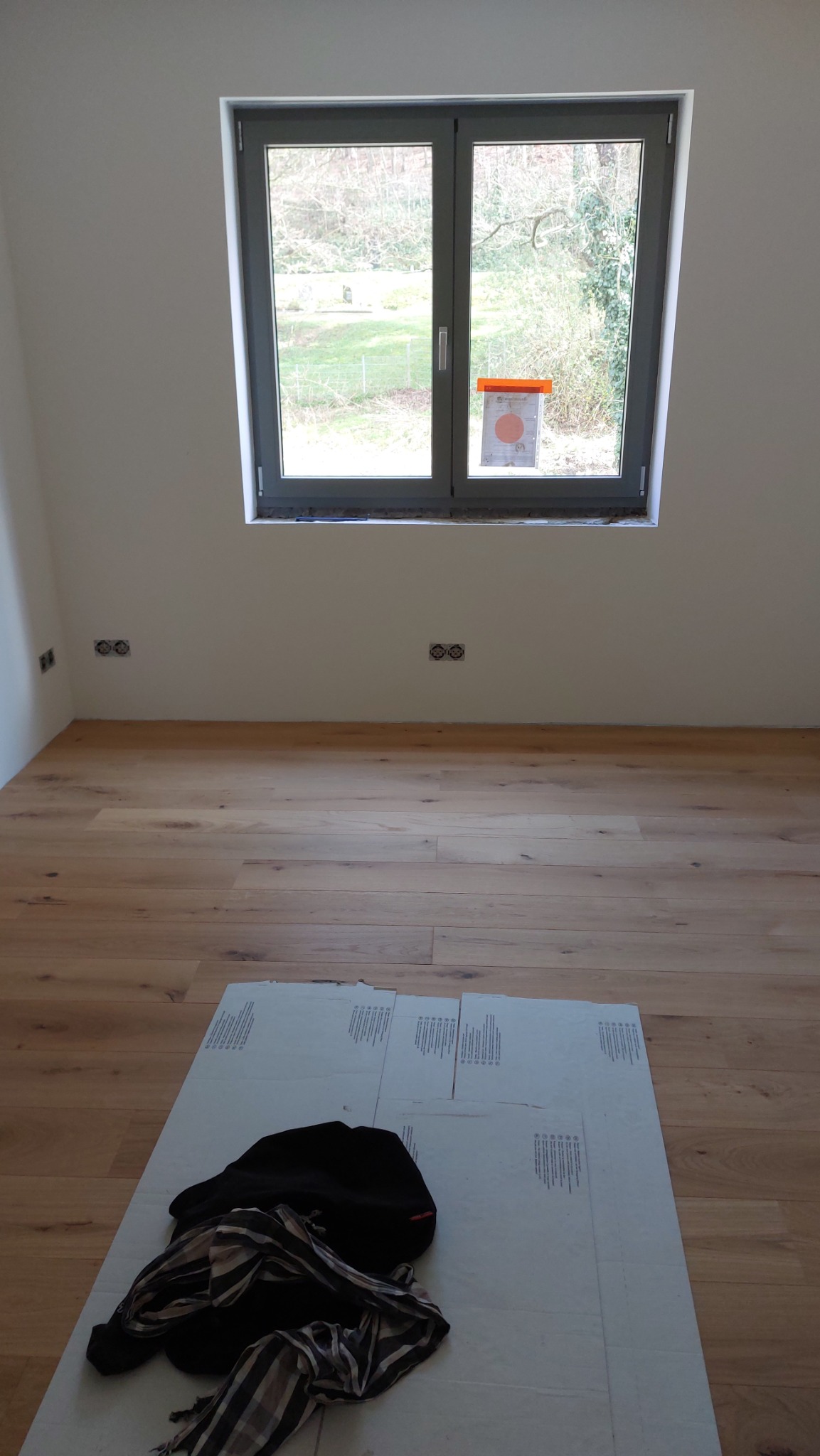 Jasna, drewniana podłoga ułożona w pustym pokoju z szarym oknem wychodzącym na zieleń. Na podłodze leżą kartony zabezpieczające i czarna bluza z szalikiem.