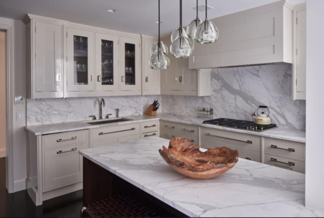 Jasna kuchnia na wymiar z marmurowymi blatami i wyspą, oświetlona szklanymi lampami wiszącymi.