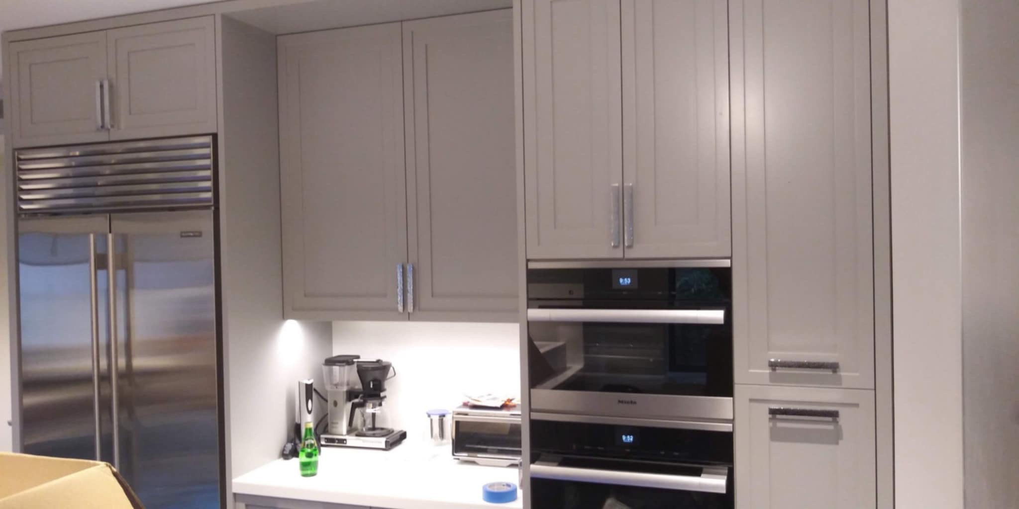 Szare szafki kuchenne z chromowanymi uchwytami, wbudowany piekarnik Miele, lodówka ze stali nierdzewnej, ekspres do kawy i toster na blacie.
