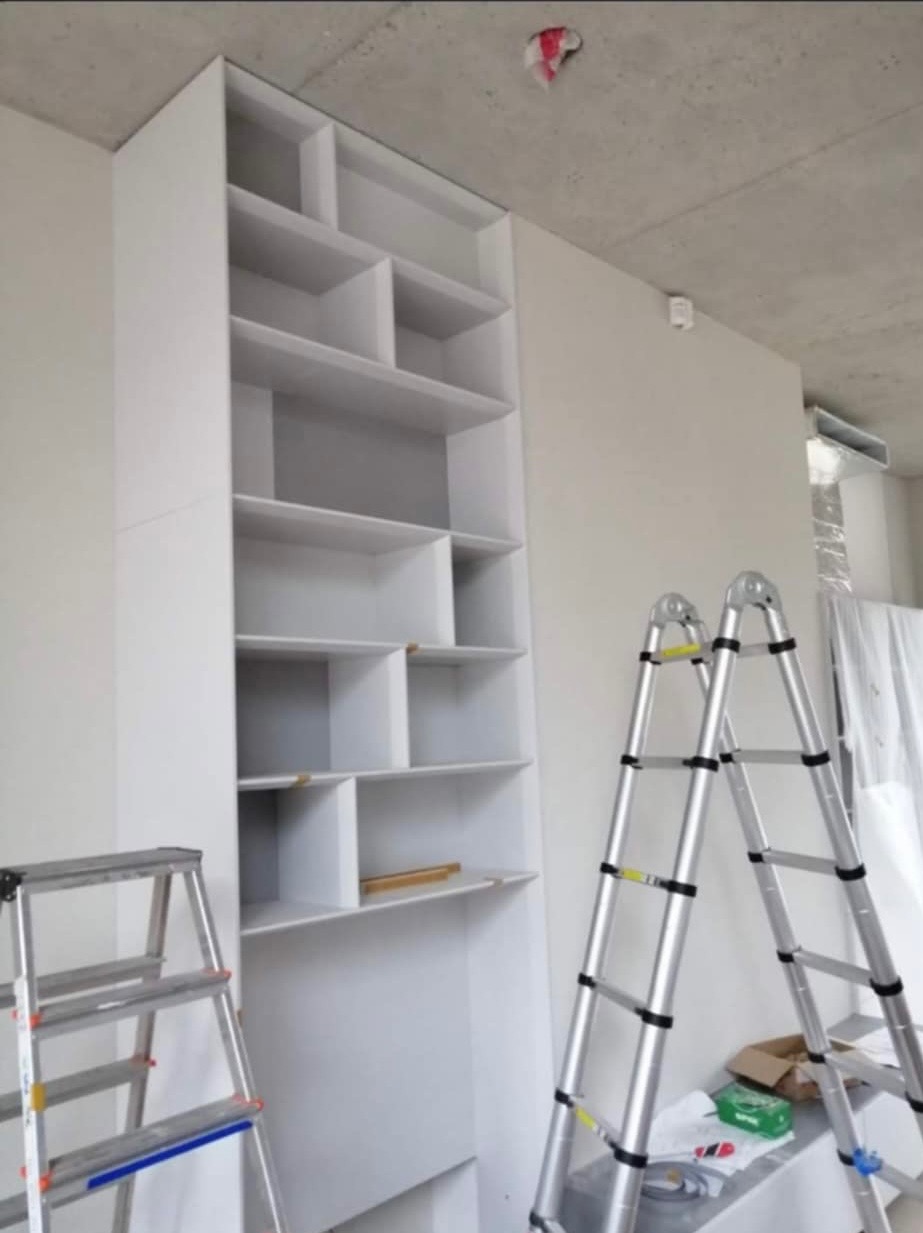 Wysoka, biała szafa wnękowa z półkami, widoczna w trakcie montażu, z dwiema rozstawionymi drabinami aluminiowymi i surowym betonowym sufitem.