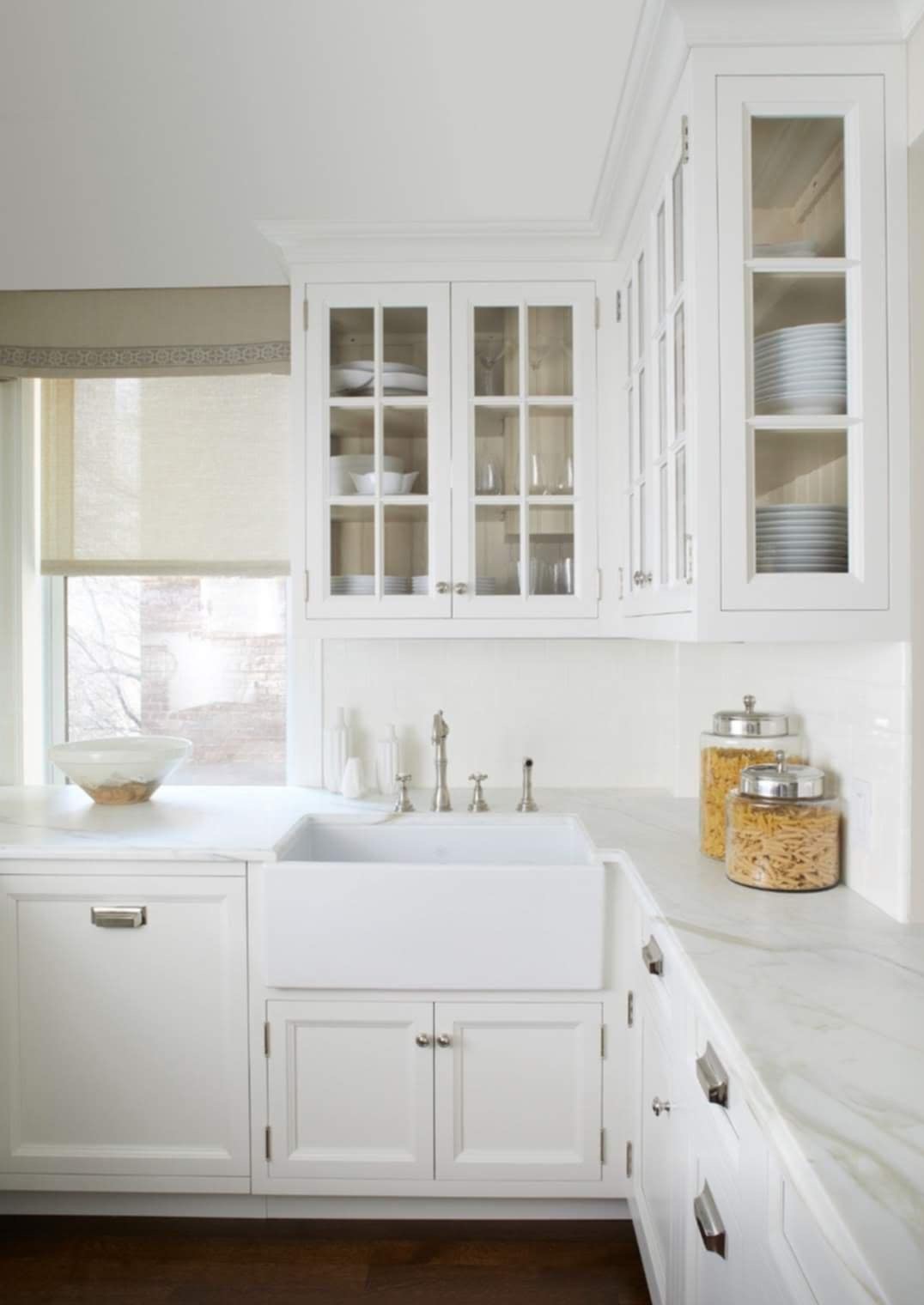 Jasna kuchnia z białymi szafkami, marmurowym blatem, ceramicznym zlewem farmerskim i przeszklonymi szafkami górnymi wypełnionymi naczyniami, z dwoma słoikami makaronu na blacie.