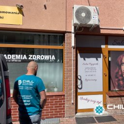 Chill Zone Mateusz Tomaszczuk - Zamontowana klimatyzacja Rotenso na budynku z salonem fryzjerskim i Akademią Zdrowia. Pracownik w koszulce Chill Zone stoi przed budynkiem.