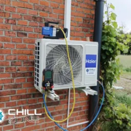 Biała klimatyzacja Haier zamontowana na ceglanej ścianie domu, podłączona do pompy próżniowej i manometru. Obok rynna z pnącą roślinnością.