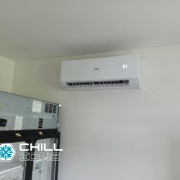 Chill Zone Mateusz Tomaszczuk - Biały klimatyzator Haier zamontowany na ścianie, wyświetlający temperaturę 21 stopni, z widoczną chłodziarką w tle. Kąt ujęcia z dołu.